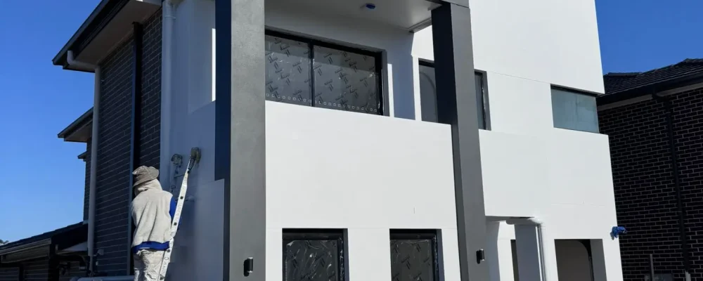 A Magic Hand Painting team member painting the exterior wall of a modern two-story house using a roller while standing on a ladder.